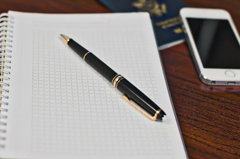 A pen on a grid notebook beside a smartphone and a passport on a wooden desk