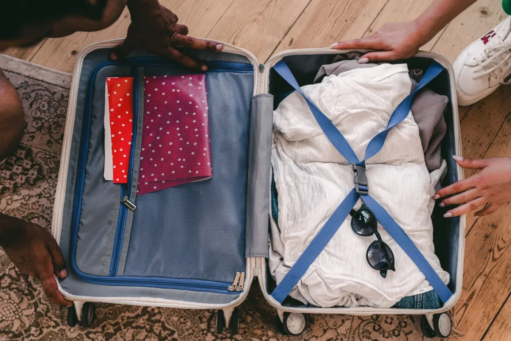 Hands around an open suitcase containing folded clothes, notebooks and sunglasses